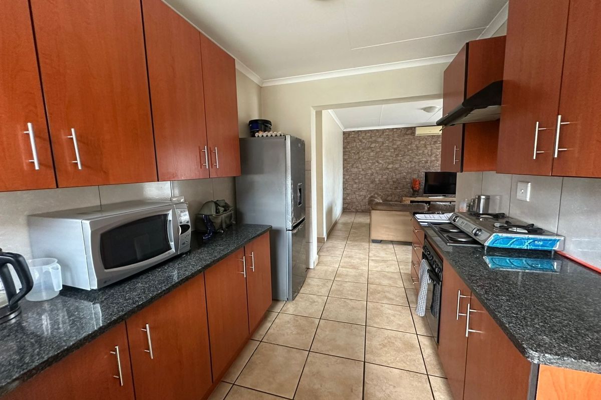 A functional kitchen featuring wood-finish cabinetry, granite countertops, a gas hob with extractor fan, and tiled flooring, leading into an adjacent living area.