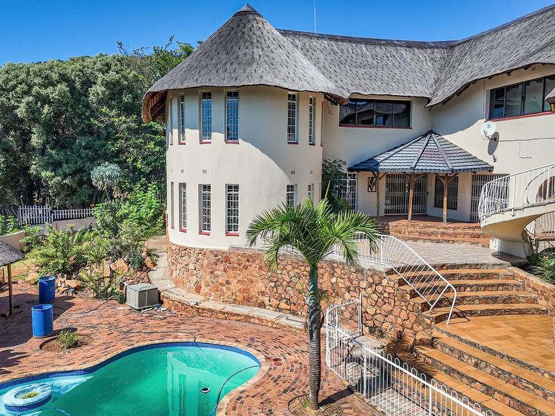 Rear exterior view featuring a swimming pool, stone-clad retaining walls, brick paved patio, and a distinctive thatch roof structure.