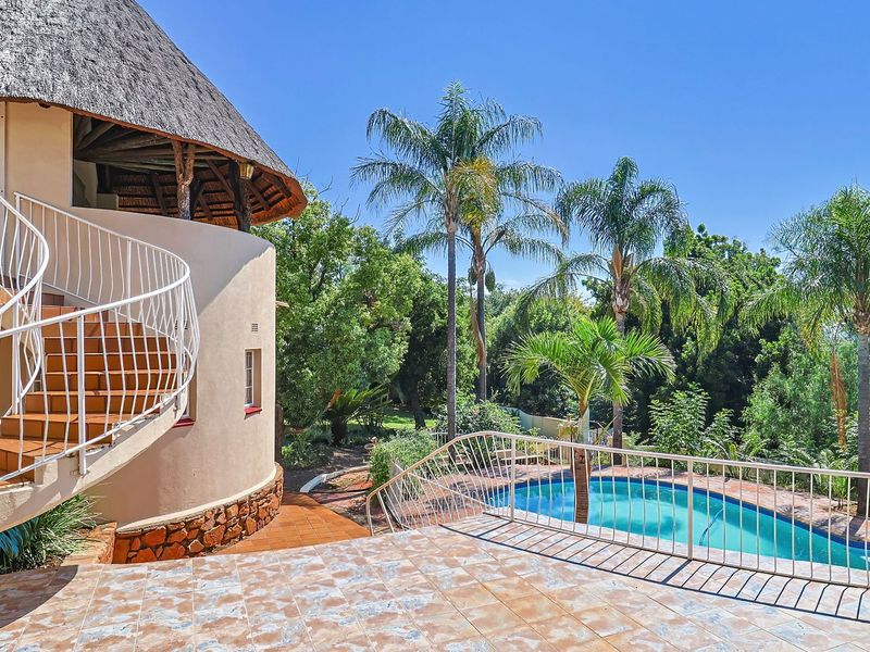 Rear exterior view featuring a thatched roof house, a tiled patio with a spiral staircase, and a swimming pool set within a landscaped garden.