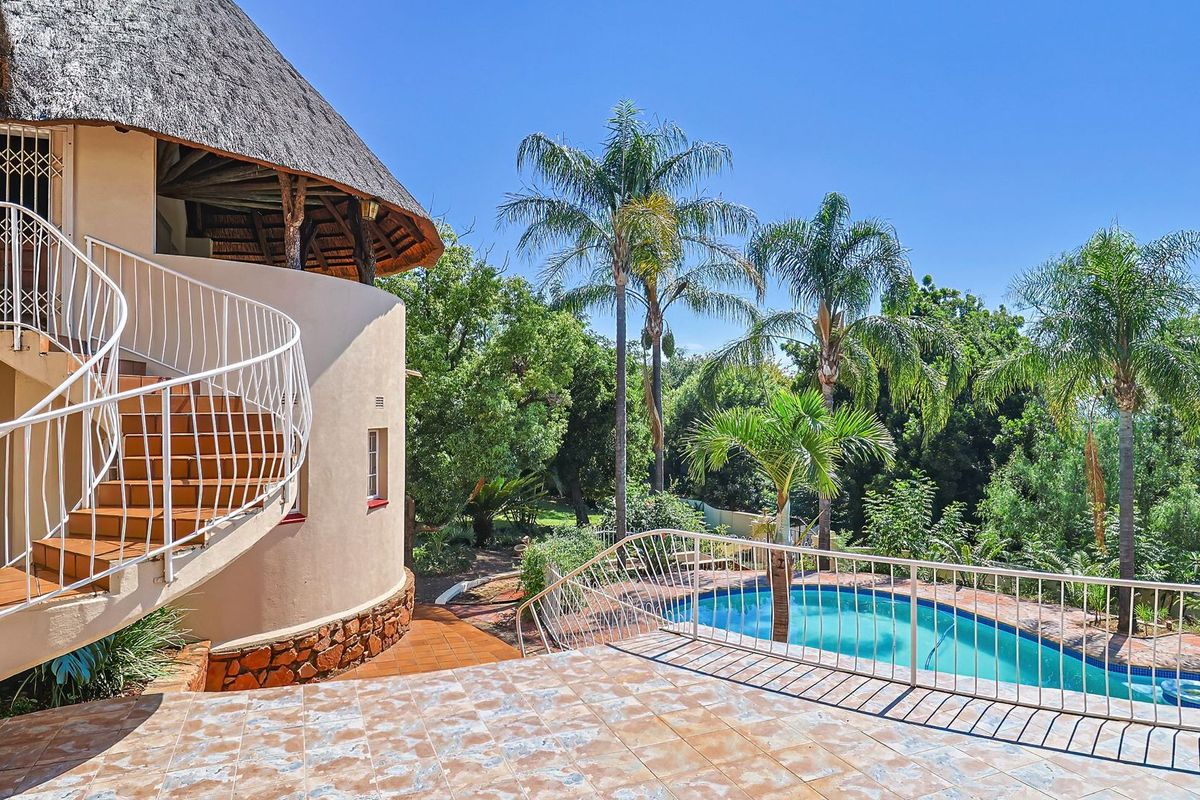 Rear exterior view featuring a thatched roof house, a tiled patio with a spiral staircase, and a swimming pool set within a landscaped garden.