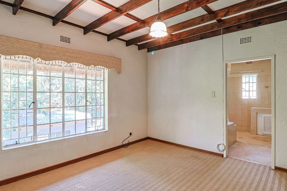 A spacious bedroom featuring exposed timber ceiling beams, carpeted flooring, and direct access to an en-suite bathroom with security bars on the window.