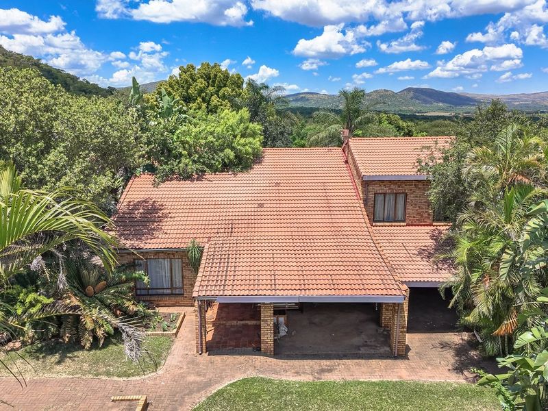 Aerial view of a brick farmhouse featuring a terracotta tile roof, covered carport, and established garden set against a scenic mountain landscape.