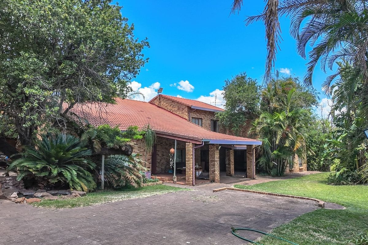 Rear view of a brick farmhouse featuring a covered patio, tiled roof, and expansive garden area with mature trees and paved driveway.