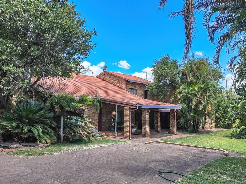 Rear view of a brick farmhouse featuring a covered patio, tiled roof, and expansive garden area with mature trees and paved driveway.