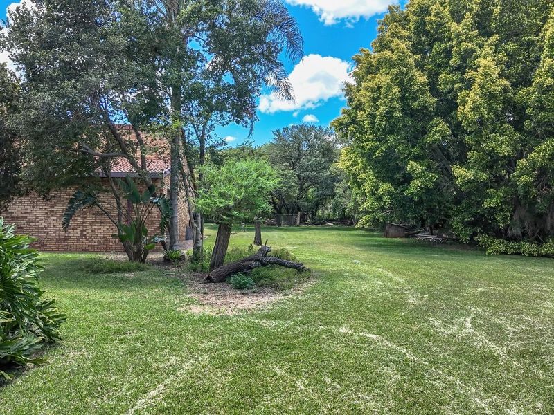 A spacious, well-established garden area featuring mature trees, lush lawn, and a view of the brick-built farm dwelling with a tiled roof.
