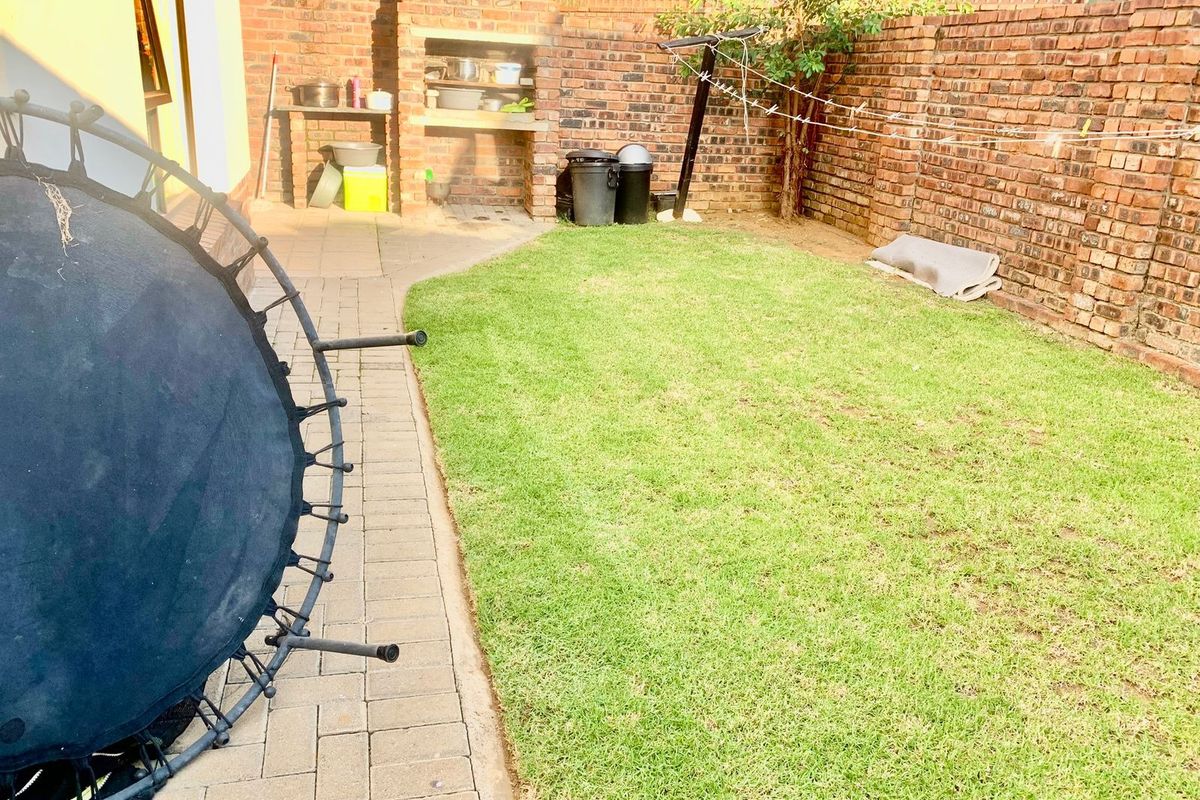 A private rear garden featuring a paved walkway, a built-in brick braai, and a well-maintained lawn enclosed by brick boundary walls.