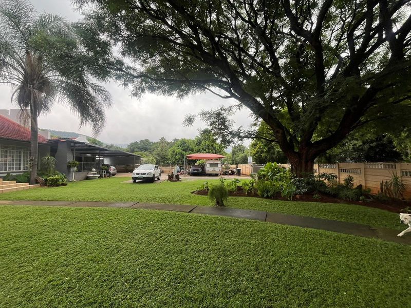 A well-maintained garden featuring a paved walkway, mature trees, and a functional carport area within a secure boundary wall.