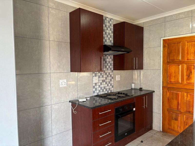 A compact kitchen featuring dark wood cabinetry, granite countertops, a built-in oven, electric hob, extractor fan, and neutral wall tiling.