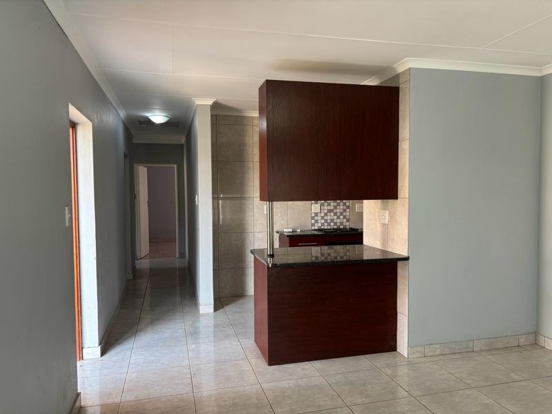 A compact kitchen area featuring dark wood cabinetry, granite countertops, a breakfast bar, and neutral tiled flooring, leading into a hallway.