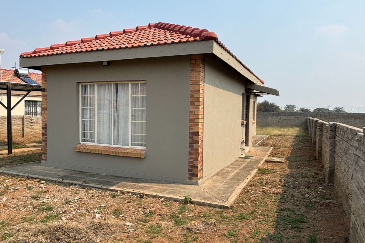 Rear view of a detached house featuring terracotta roof tiles, brick corner accents, and a concrete apron surrounding the structure within a fenced yard.