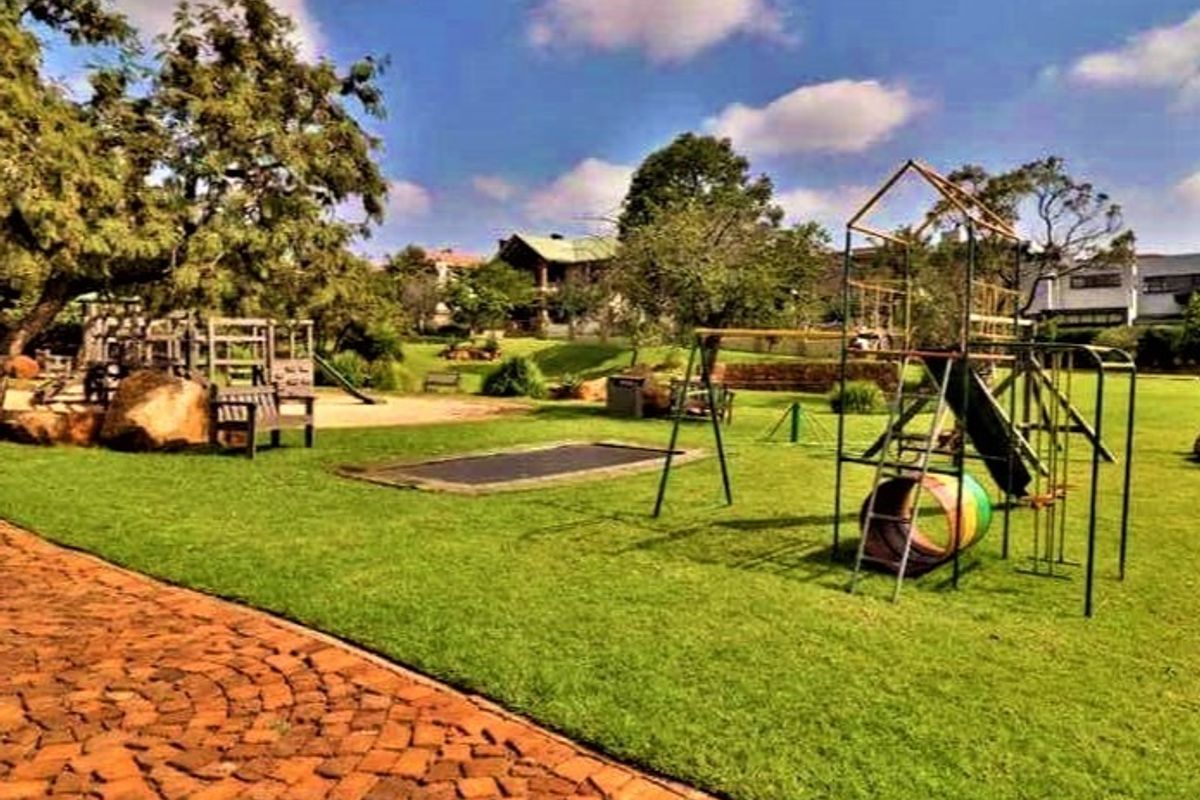 A well-maintained residential garden featuring a large lawn, mature trees, paved pathways, and integrated playground equipment for recreational use.