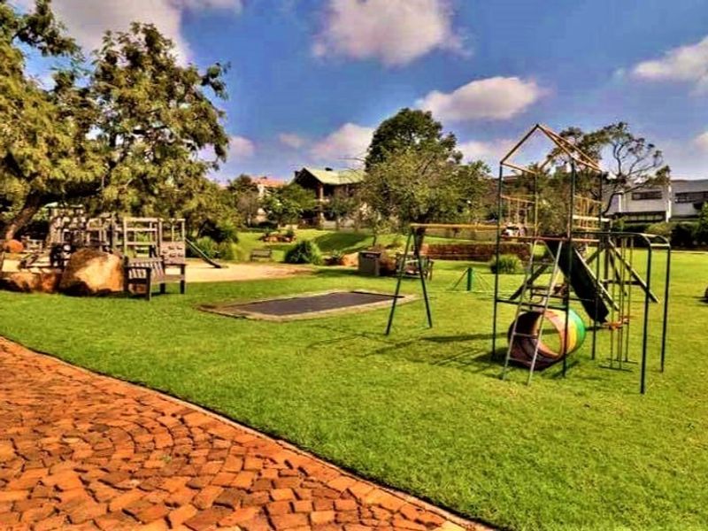 A well-maintained residential garden featuring a large lawn, mature trees, paved pathways, and integrated playground equipment for recreational use.