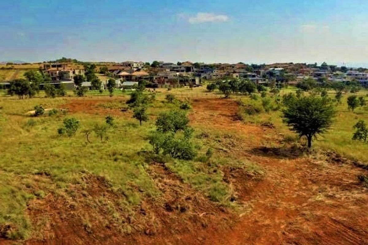 An aerial view of a vacant residential plot featuring natural terrain and vegetation with surrounding neighborhood development in the background.