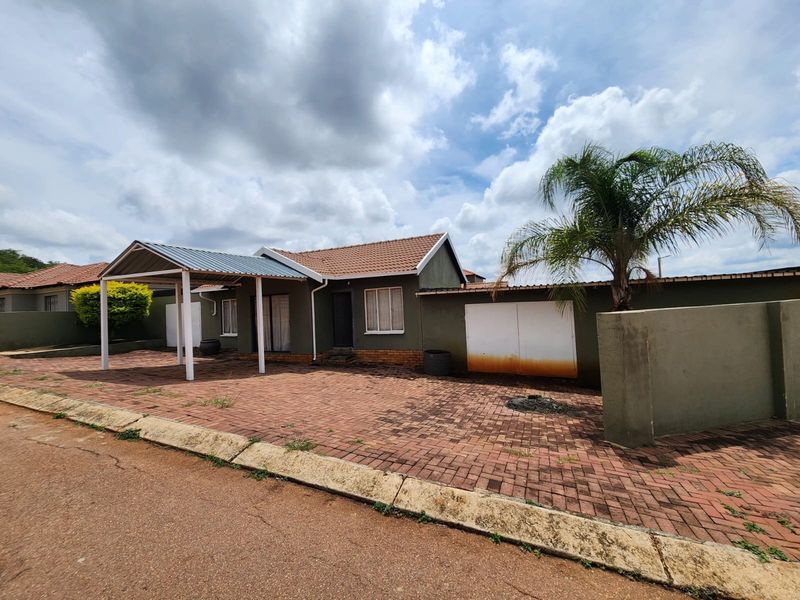 Front exterior view of a house featuring a paved driveway, a covered carport, and a boundary wall with a metal garage door.