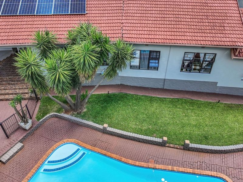 Aerial view of the property featuring a swimming pool, paved patio area, landscaped lawn, and a tiled roof equipped with solar panels.