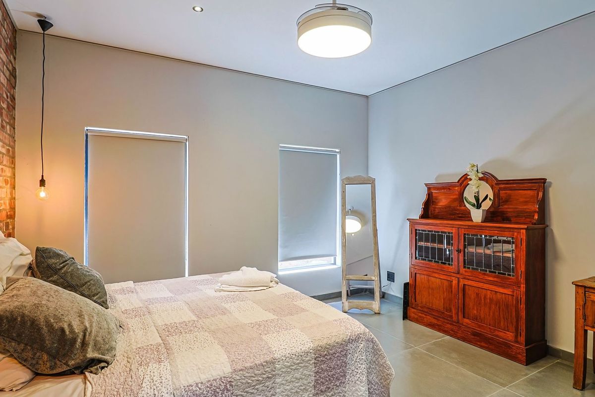 A well-maintained bedroom featuring an exposed brick accent wall, neutral tiled flooring, and modern recessed lighting with roller blinds for privacy.