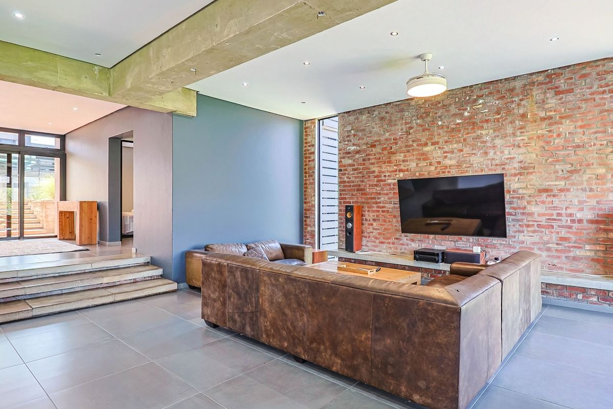 Spacious living room featuring an industrial aesthetic with an exposed brick feature wall, concrete beam, and tiled flooring leading to an adjacent area.
