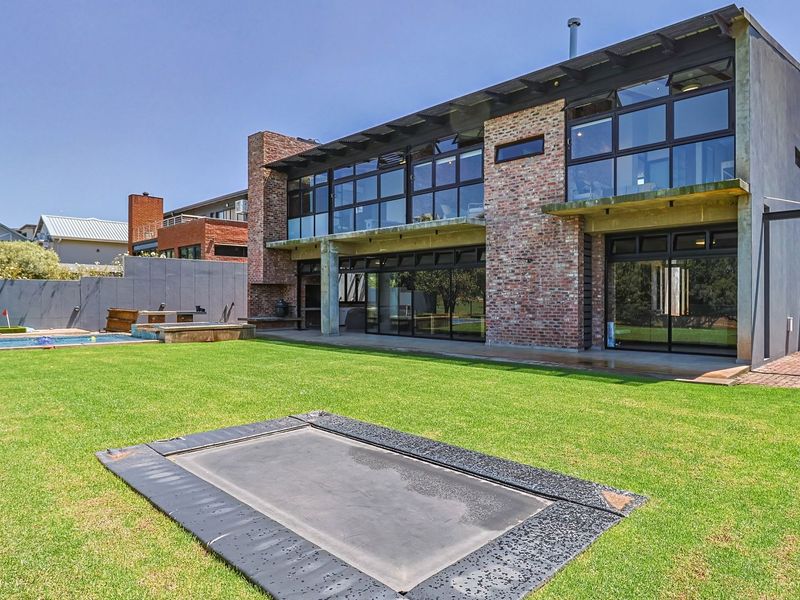 Rear view of a modern two-story home featuring exposed brickwork, extensive glazing, a concrete balcony, and a well-maintained garden with a swimming pool.