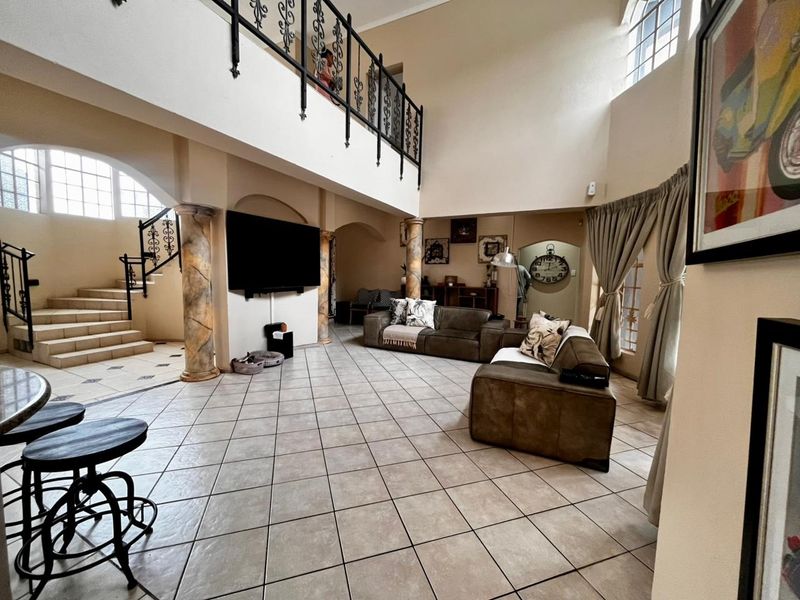 Spacious double-volume living area featuring tiled floors, decorative columns, a wrought iron staircase, and a mezzanine level overlooking the main lounge space.