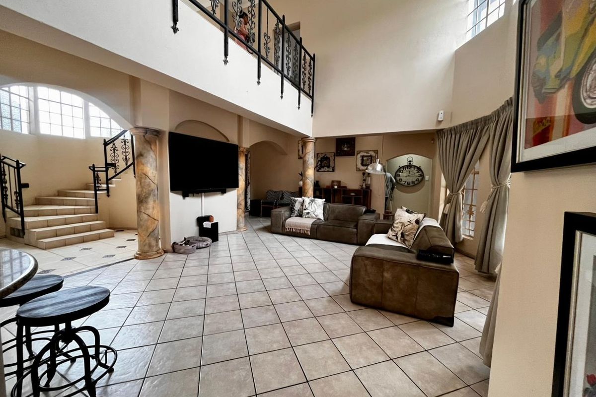 Spacious double-volume living area featuring tiled floors, decorative columns, a wrought iron staircase, and a mezzanine level overlooking the main lounge space.