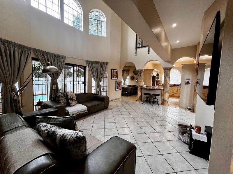 Spacious open-plan living area featuring double-volume ceilings, large arched windows, tiled flooring, and decorative columns leading into a functional kitchen space.