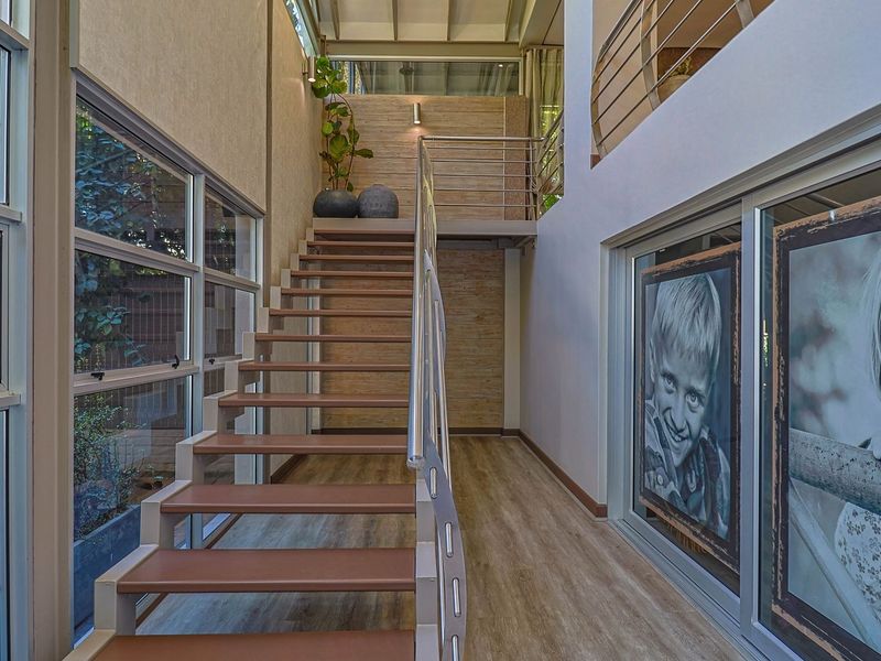 A bright, modern double-volume hallway featuring a contemporary staircase with metal balustrades, large glass windows, and laminate flooring leading to a mezzanine level.
