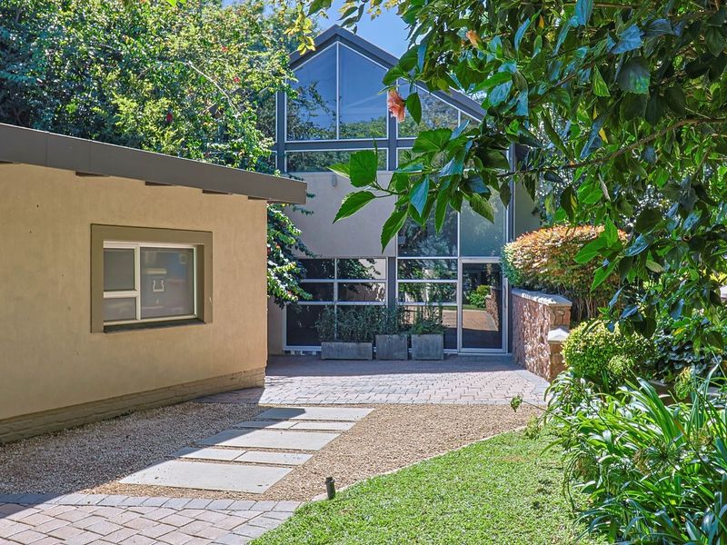 A well-maintained rear exterior featuring a paved patio, stone retaining wall, and a striking modern glass facade overlooking a landscaped garden.