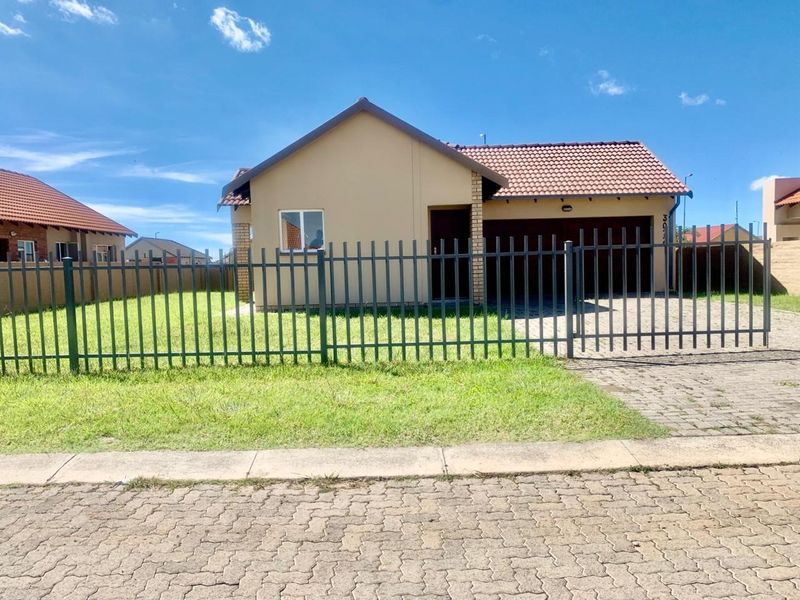 Front view of a single-story residential house featuring a paved driveway, garage, and secure metal perimeter fencing.