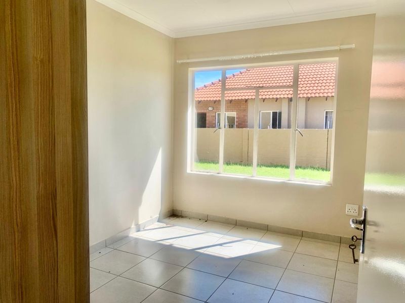 A bright, empty bedroom featuring neutral tiled flooring, a large window providing natural light, and a built-in cupboard.