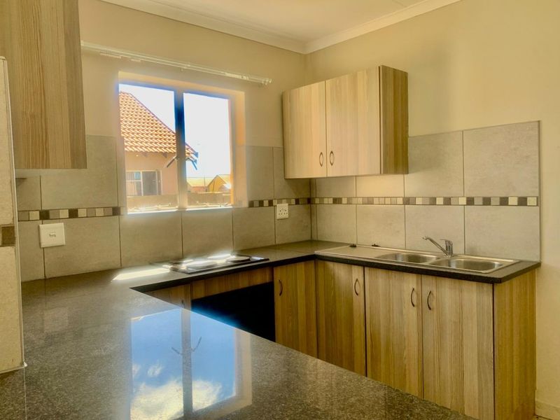 A clean, modern kitchen featuring light wood-finish cabinetry, dark stone-look countertops, a built-in electric hob, and a double bowl sink with tiled backsplash.