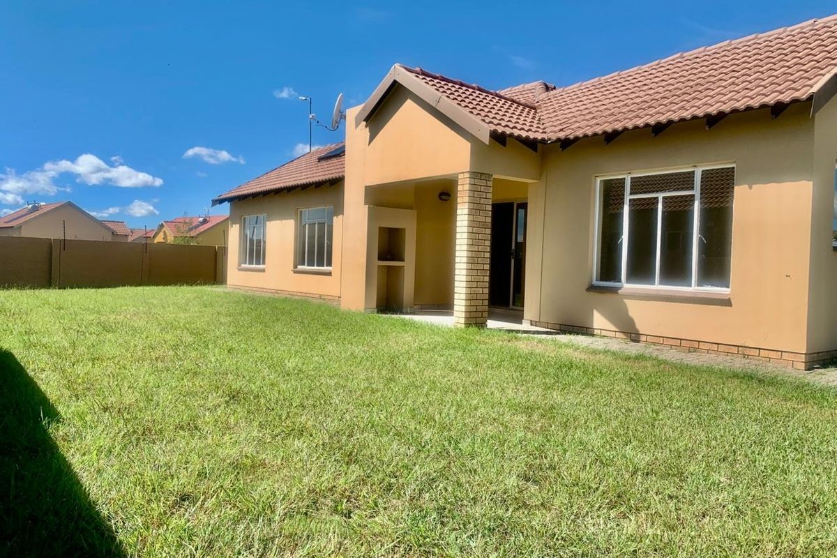 Rear view of the house featuring a tiled roof, brick facade, covered patio with a built-in braai, and a spacious lawn area.