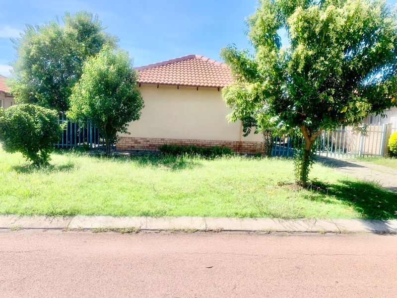A view of the front exterior of the house featuring a tiled roof, brick plinth, and metal perimeter fencing with an overgrown lawn.