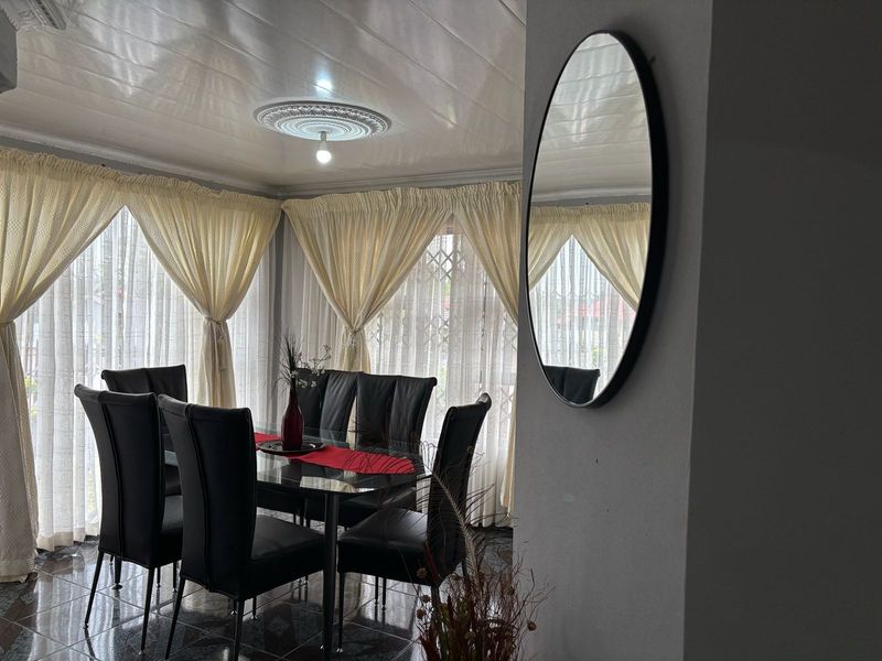 A dining room featuring tiled flooring, a decorative ceiling rose, and large windows with curtains, providing a bright and functional space for meals.