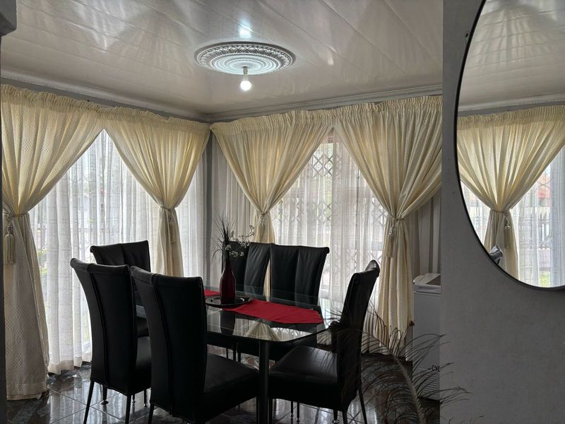 A dining room featuring tiled flooring, a decorative ceiling rose, and large windows covered by curtains, providing a bright and functional space.