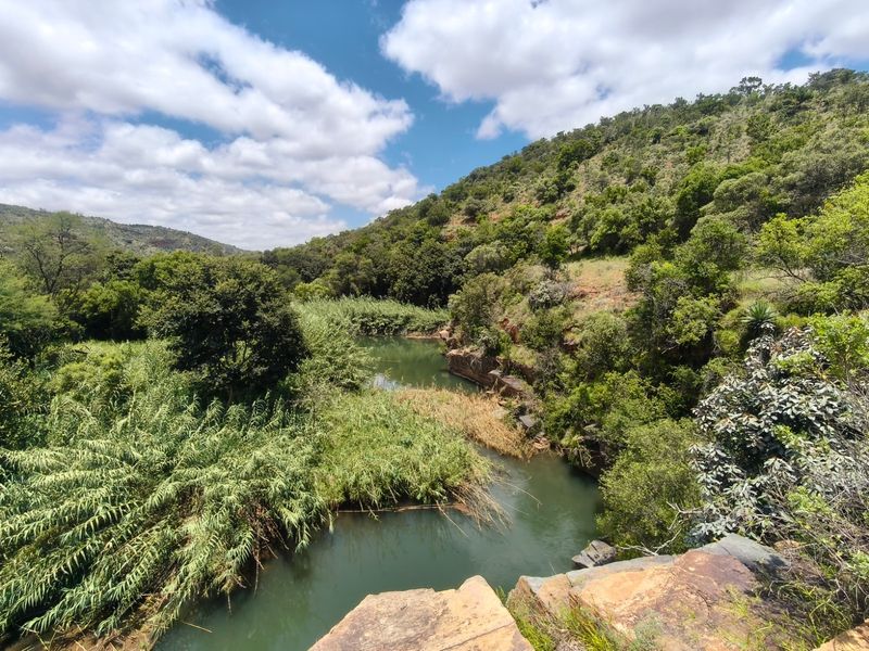 A scenic view of a natural river flowing through a lush, hilly landscape with rocky outcrops and dense vegetation.