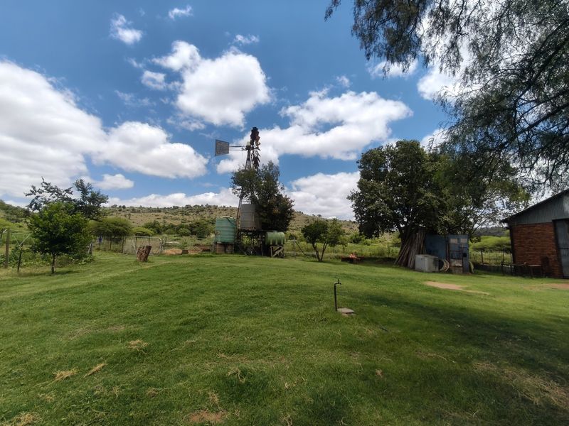 Expansive farm garden featuring a traditional windmill, water storage tanks, and an outbuilding set against a scenic rural landscape.