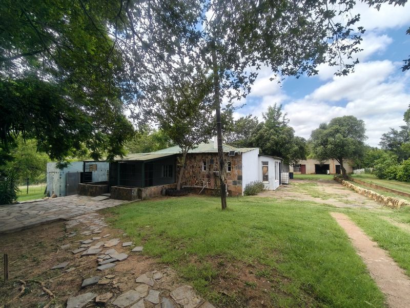 Rear view of a farm property featuring stone-clad walls, a metal roof, and an expansive garden area with paved pathways and an adjacent outbuilding.