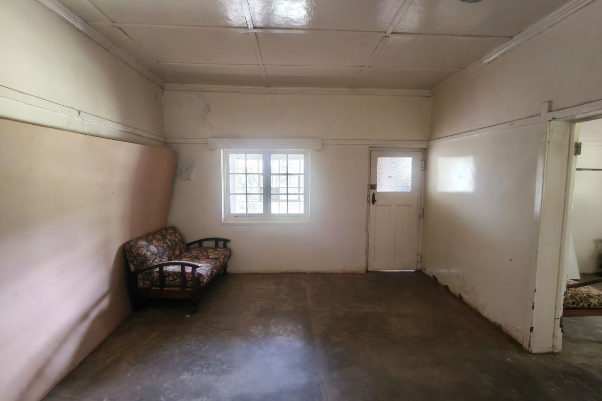 A dated living area featuring concrete floors, a timber-framed window, and a panelled door, requiring significant cosmetic refurbishment and wall repairs.