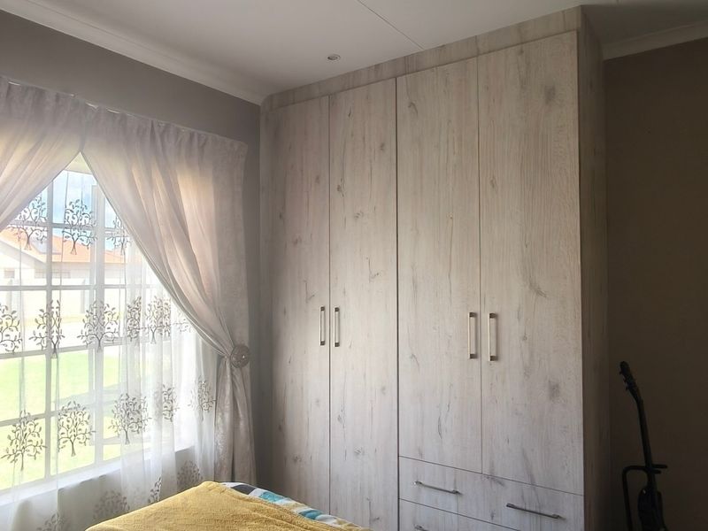 A well-maintained bedroom featuring light wood-finish built-in cupboards and a window with security bars, providing ample storage and natural light.