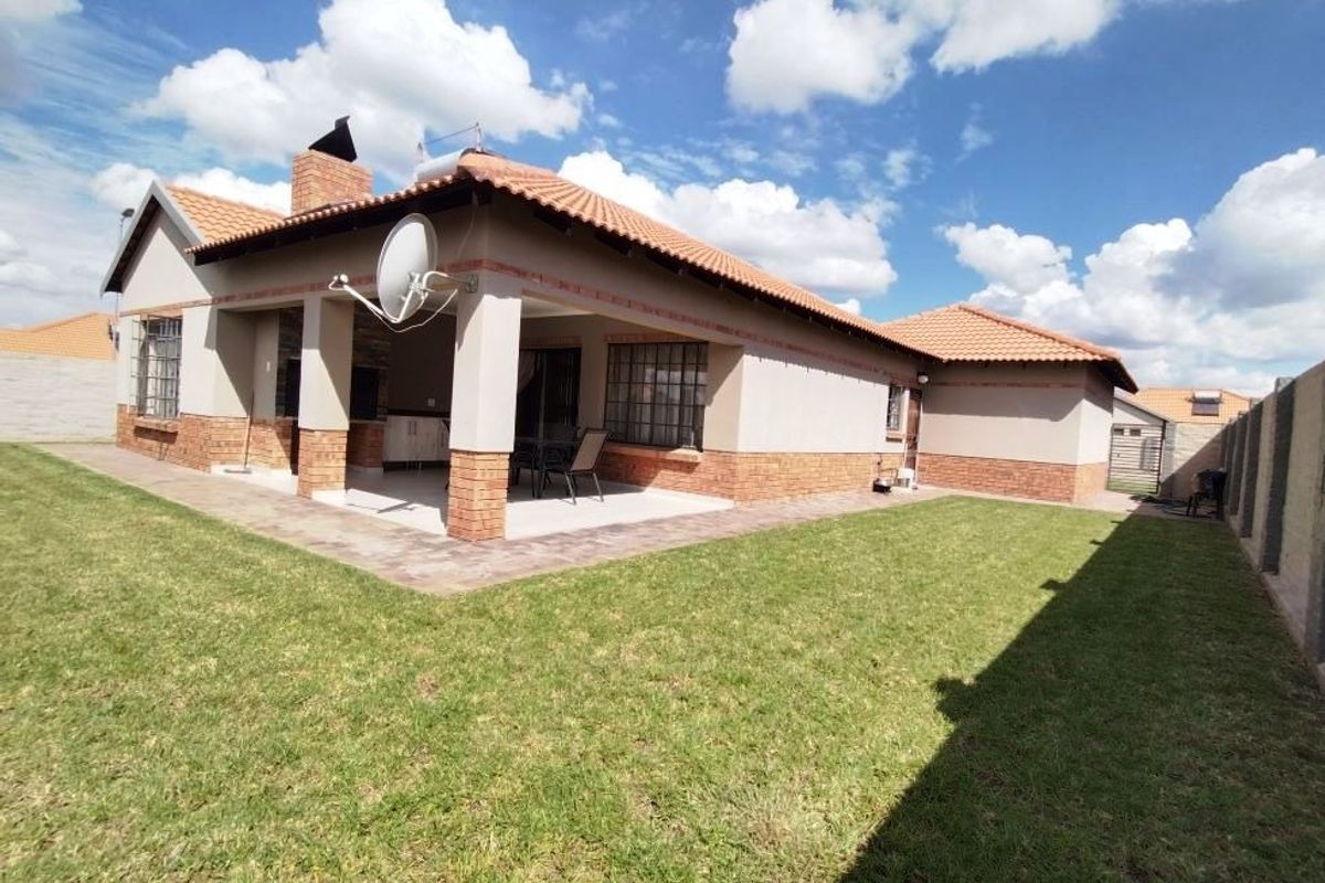 Rear view of the house featuring a covered patio, brick accents, tiled roof, and a well-maintained lawn with perimeter walling.