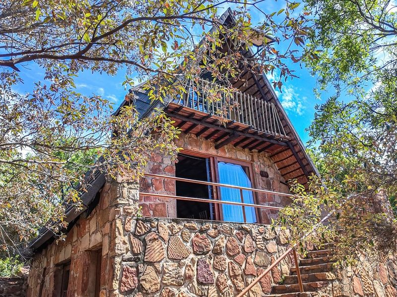 A charming A-frame house featuring rustic stone cladding, a timber balcony, and an external staircase set within a natural, wooded environment.