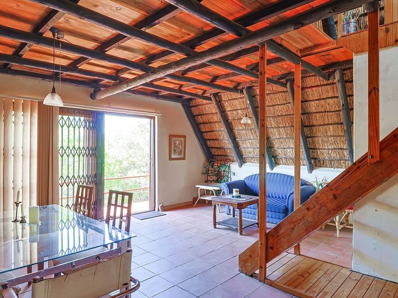 Open-plan living and dining area featuring a rustic thatched roof, exposed timber beams, tiled floors, and a wooden staircase leading to an upper level.