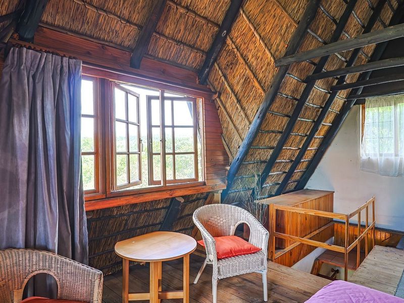 A rustic loft-style bedroom featuring exposed timber rafters, a traditional thatched roof ceiling, and timber casement windows overlooking the surrounding landscape.