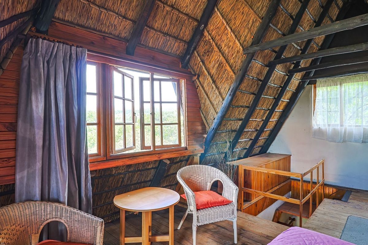 A rustic loft-style bedroom featuring exposed timber rafters, a traditional thatched roof ceiling, and timber casement windows overlooking the surrounding landscape.