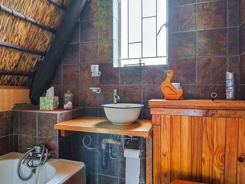 A rustic bathroom featuring a thatched roof with exposed timber rafters, dark tiled walls, a vessel sink on a timber vanity, and a bathtub.