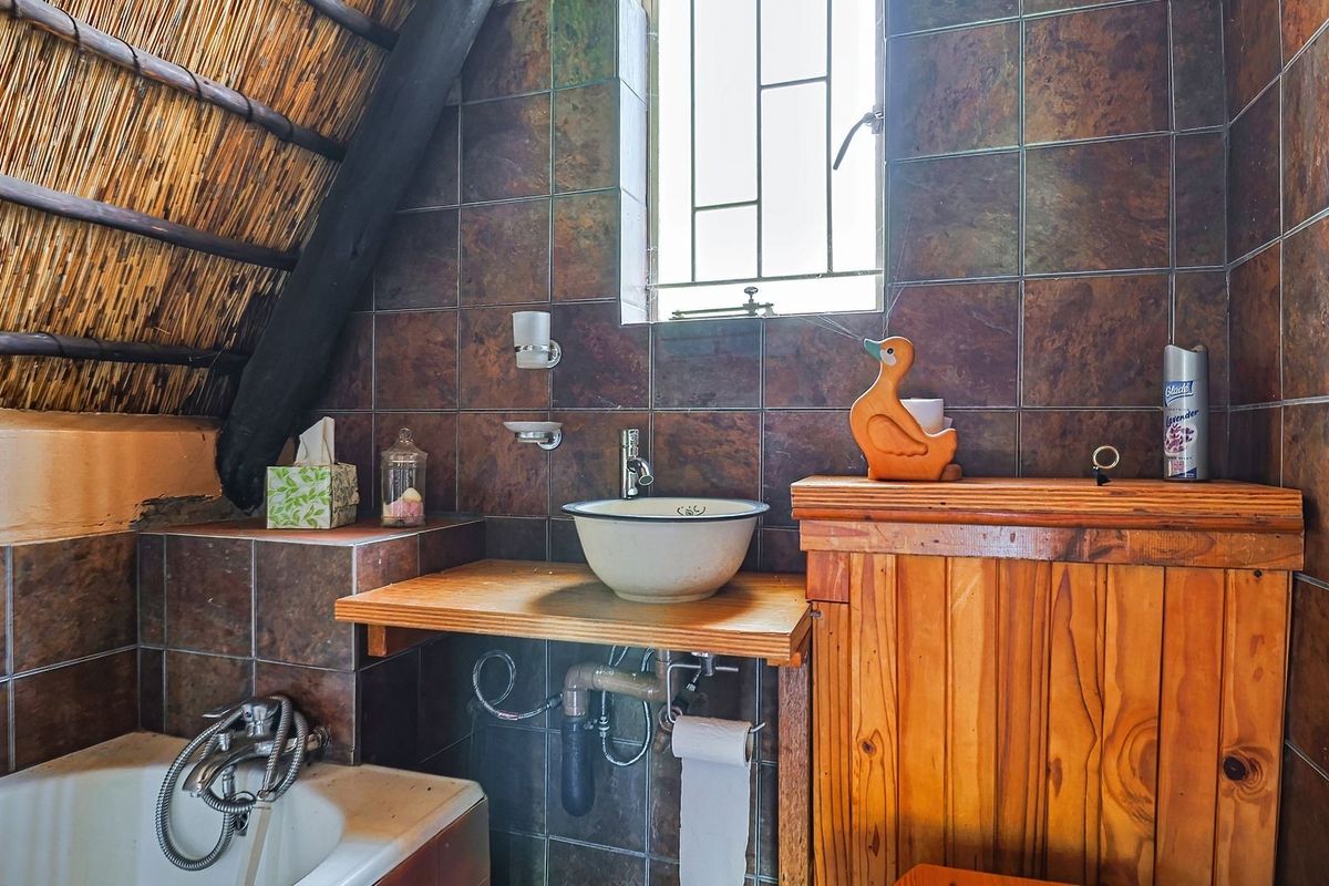 A rustic bathroom featuring a thatched roof with exposed timber rafters, dark tiled walls, a vessel sink on a timber vanity, and a bathtub.