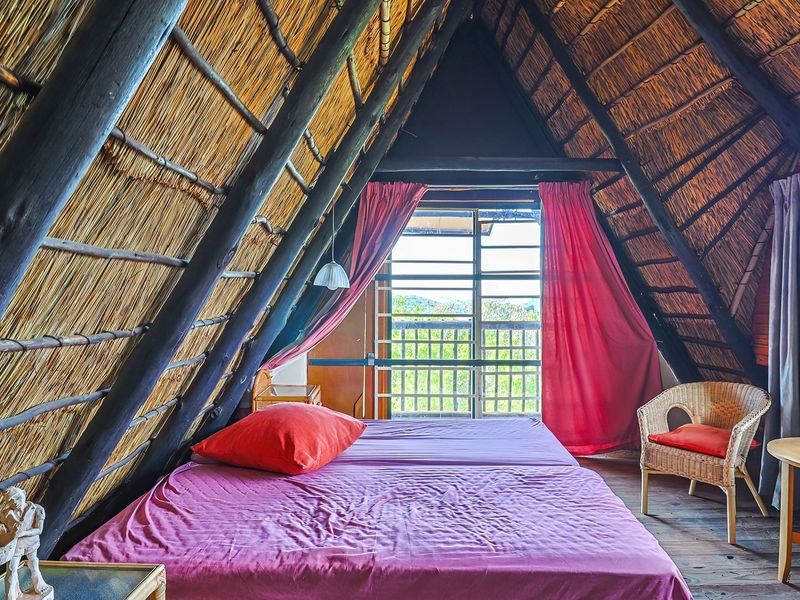 A rustic bedroom featuring a distinctive A-frame design with exposed timber rafters, a thatched roof, and direct access to a balcony through sliding doors.