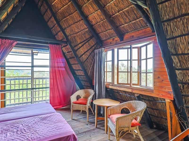 A rustic bedroom featuring exposed timber rafters, a traditional thatched roof interior, and timber flooring with views through large casement windows.