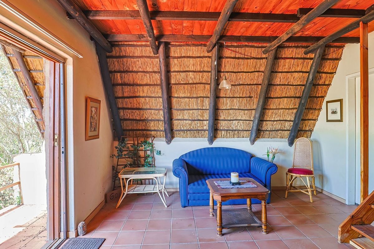 A rustic living area featuring a distinctive thatched roof with exposed timber rafters, tiled flooring, and direct access to an outdoor balcony.