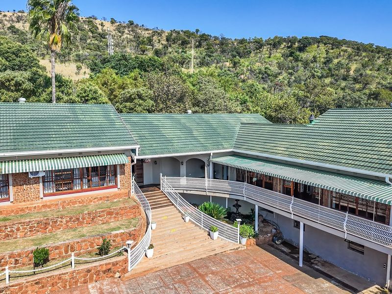 Elevated view of a farm-style residence featuring stone cladding, green tiled roofing, expansive paved courtyard, and tiered retaining walls set against a hillside.